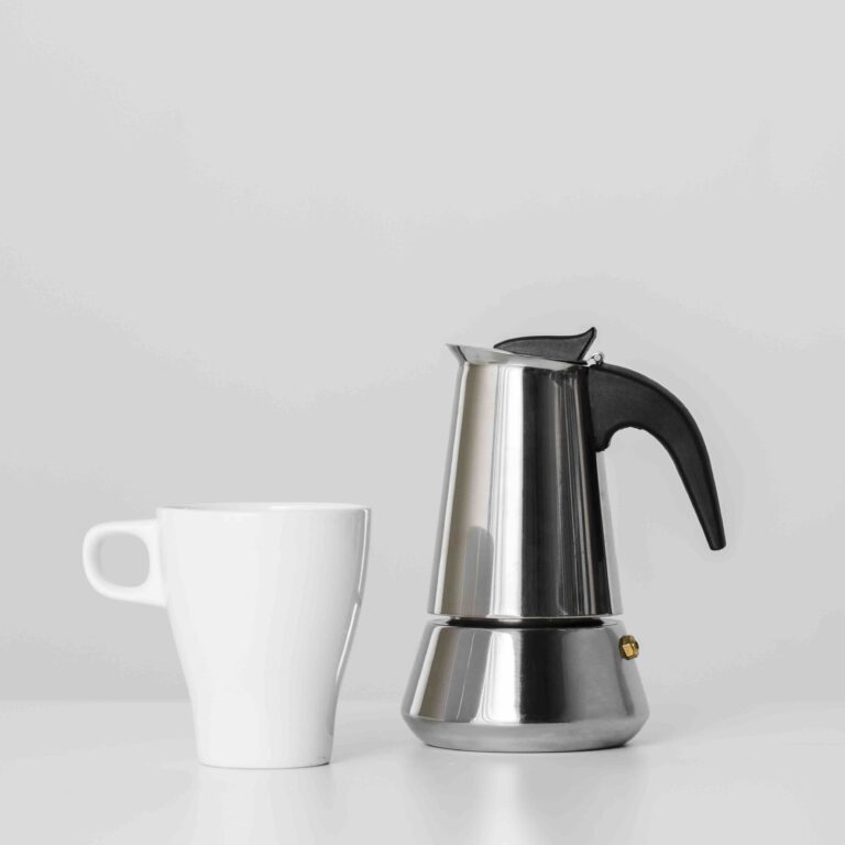 Moka in acciaio inox con tazza bianca su sfondo minimal, confronto tra caffettiere in acciaio e alluminio