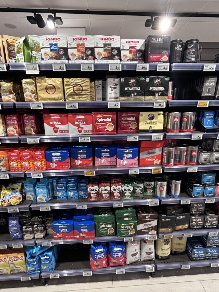 Scaffale supermercato con confezioni di caffè commerciali italiani: Lavazza, Kimbo, Illy e altre marche